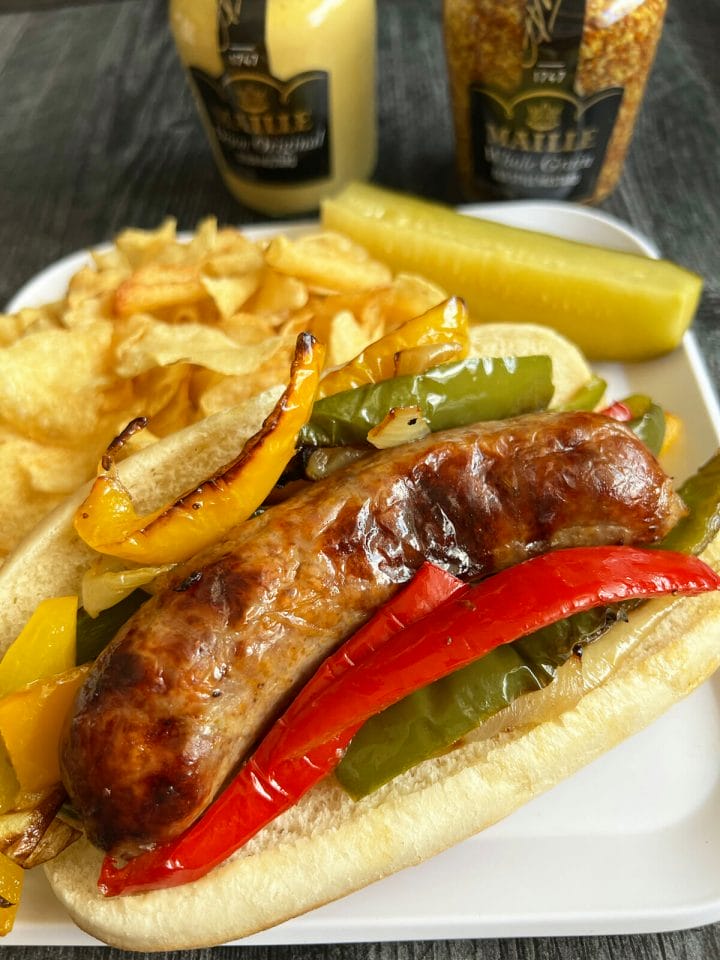 A plate with a sausage and peppers sandwich, chops and a pickle.