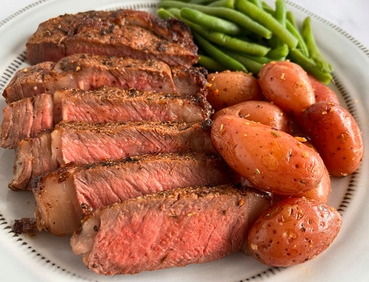A plate of sliced ribeye steak made in an air fryer, served with potatoes and green beans.