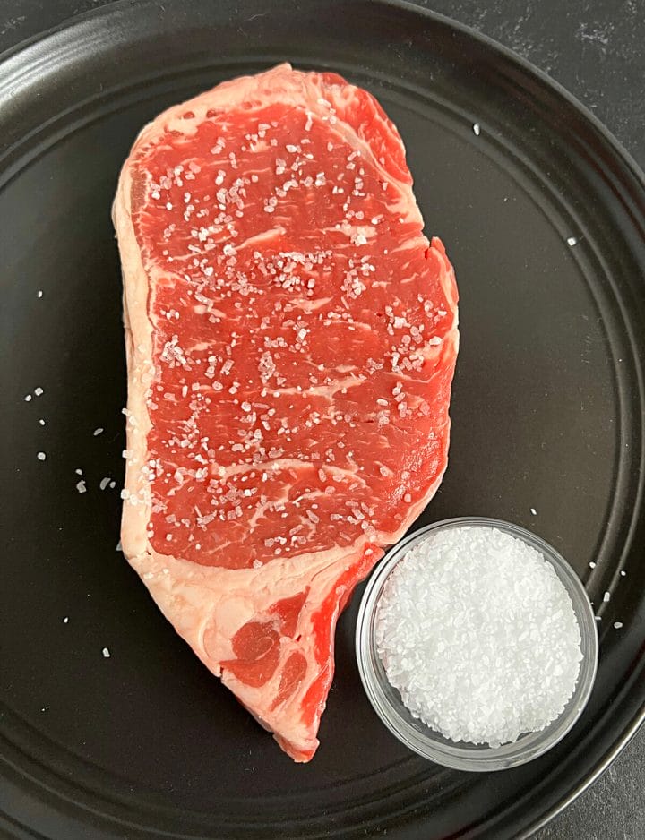 A New York strip steak being seasoned with kosher salt.