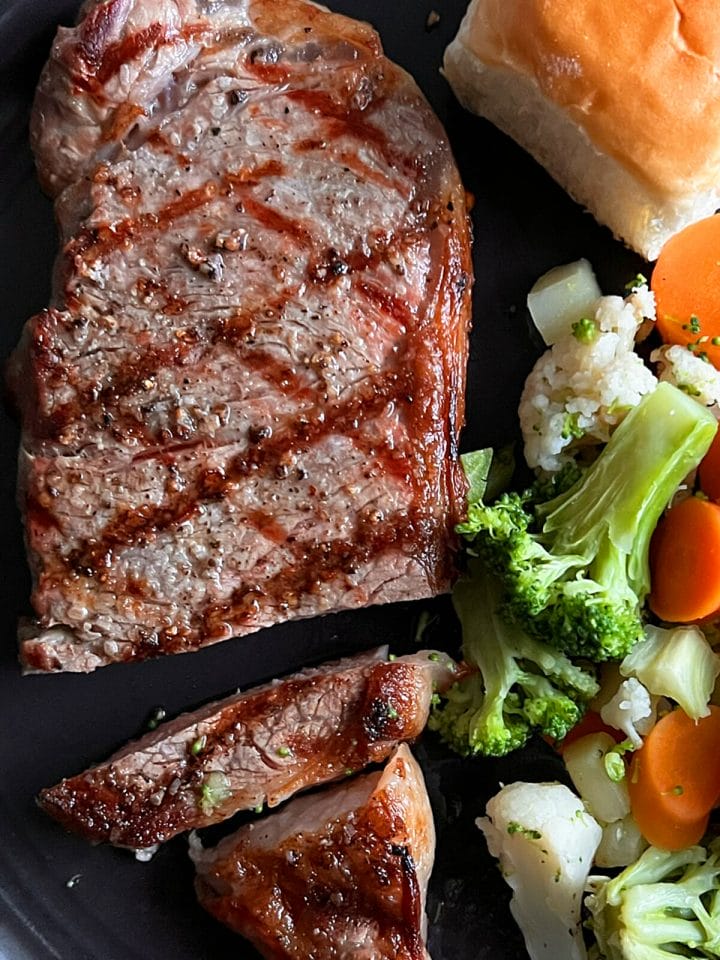 A New York strip steak seasoned with kosher salt, pepper, and served with vegetables and a roll.