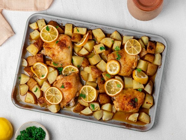 A sheet pan of oven roasted chicken with potatoes in a lemon sauce with sliced lemons and garnished with parsley.