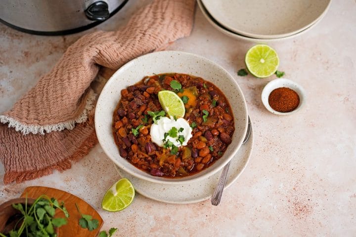 A bowl of chili made in a slow cooker garnished with sour cream, limes and parsley.