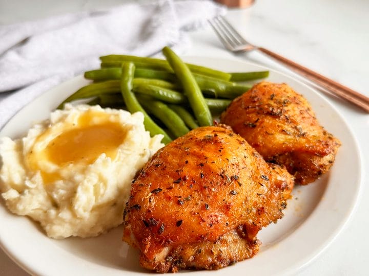 Oven baked chicken thighs served on a plate with mashed potatoes and gravy, and fresh green beans. 