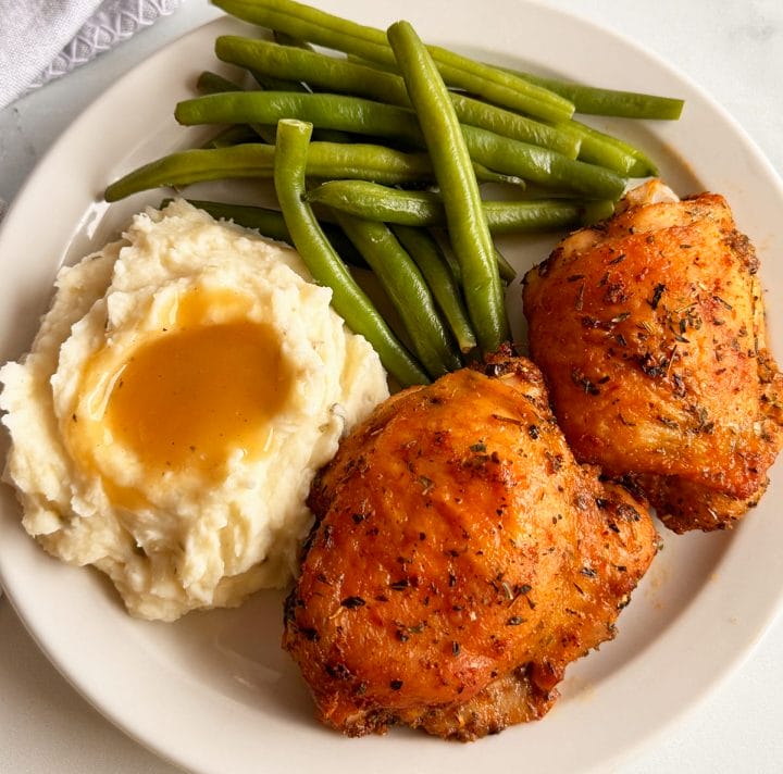 Oven baked chicken thighs served on a plate with green beans, mashed potatoes and gravy.