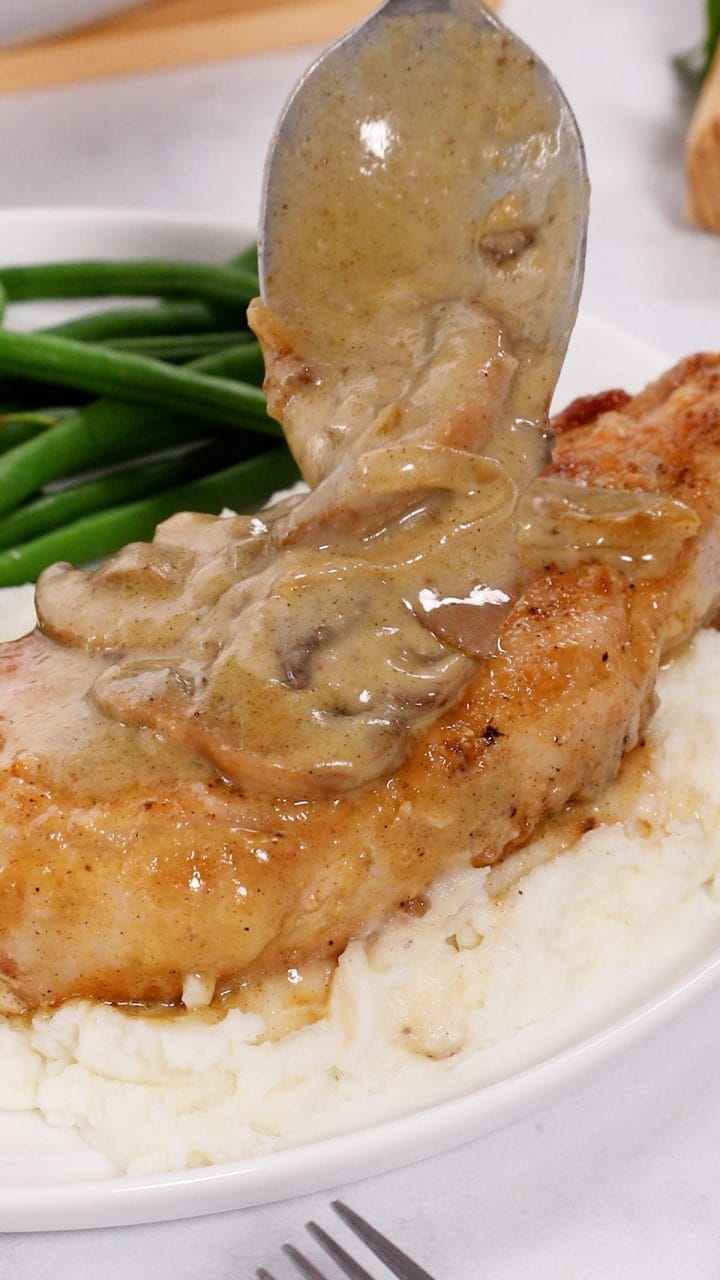 A pork chop atop mashed potatoes being topped with an onion and mushroom gravy for smothered pork chops. 